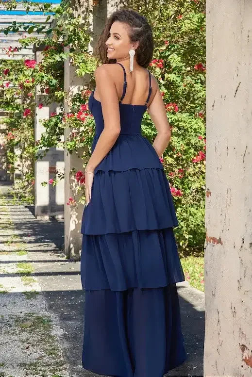 A woman wearing a navy blue, tiered gown stands smiling in a garden path adorned with vibrant pink flowers and stone columns, creating a picturesque and serene atmosphere.