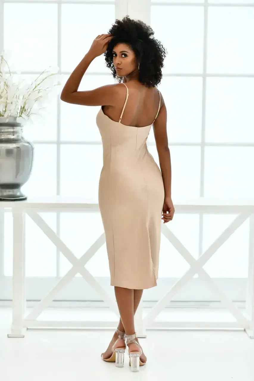 A woman in a fitted, beige dress stands with her back to the camera, turning slightly to look over her shoulder, against a bright backdrop of windows and decorative flowers.