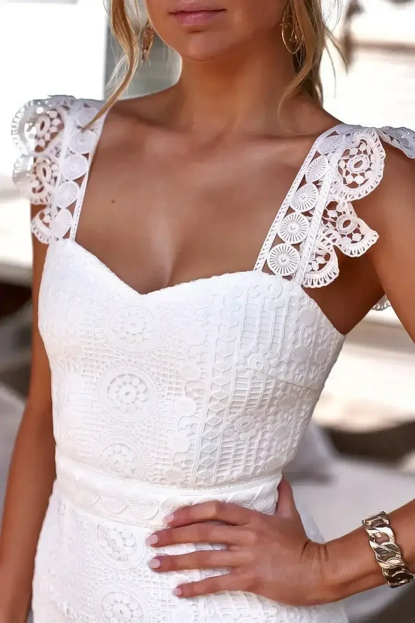 A white dress with intricately patterned fabric and lace-trimmed straps is worn by a woman resting her hand on her hip, against a softly blurred outdoor background.