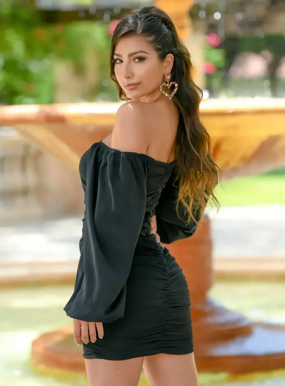 A woman poses confidently by a fountain, wearing a stylish black off-shoulder dress and vibrant, heart-shaped earrings. Lush greenery surrounds the scene, enhancing the vibrant atmosphere.