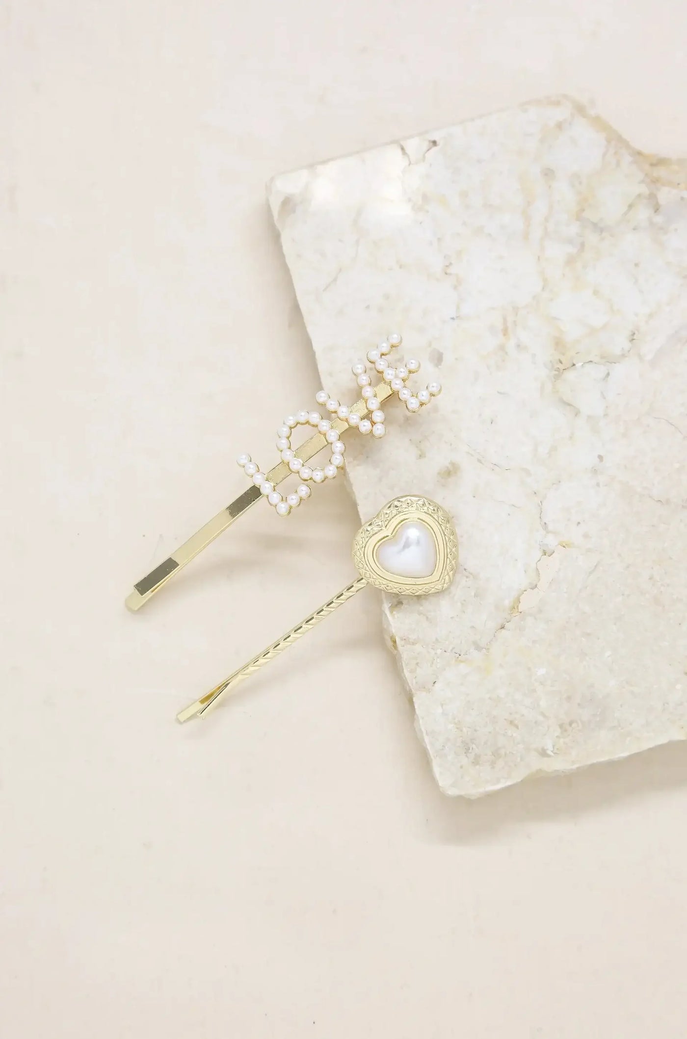 Two decorative hairpins are resting on a light-colored stone surface. One pin spells "LOVE" with small pearls; the other is heart-shaped with a pearl center.