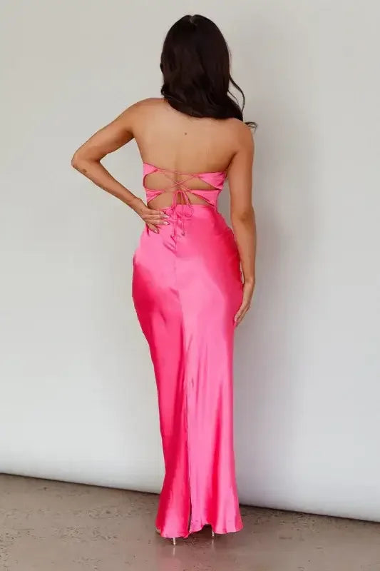 A person wearing a long, pink satin dress stands with their back facing the viewer. The dress has a strapless design and ties intricately at the back, displaying a smooth, minimalist background.