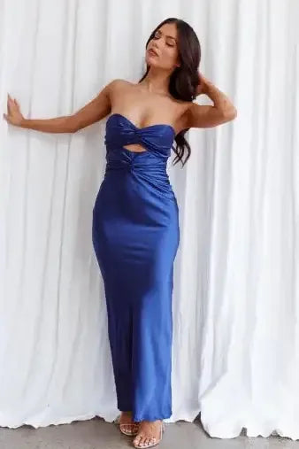 A woman in a blue satin dress poses elegantly against a white backdrop, with one hand raised and her head tilted, showcasing the dress's design and her graceful demeanor.