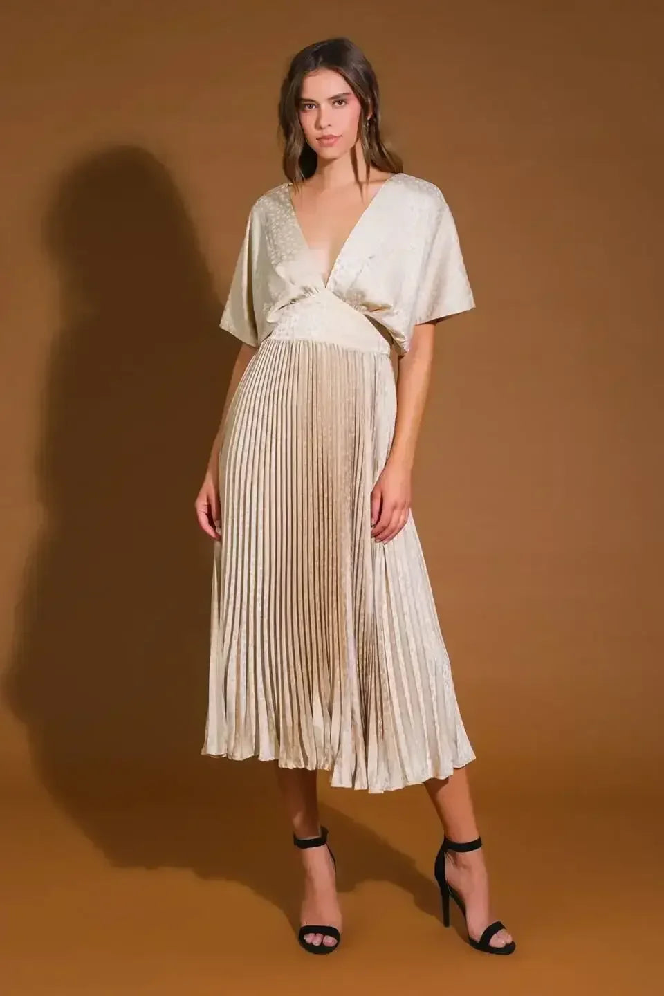 A woman stands confidently in a pleated, light-colored dress with a deep neckline and short sleeves, complemented by black high-heeled sandals. The backdrop is a warm, brown tone.