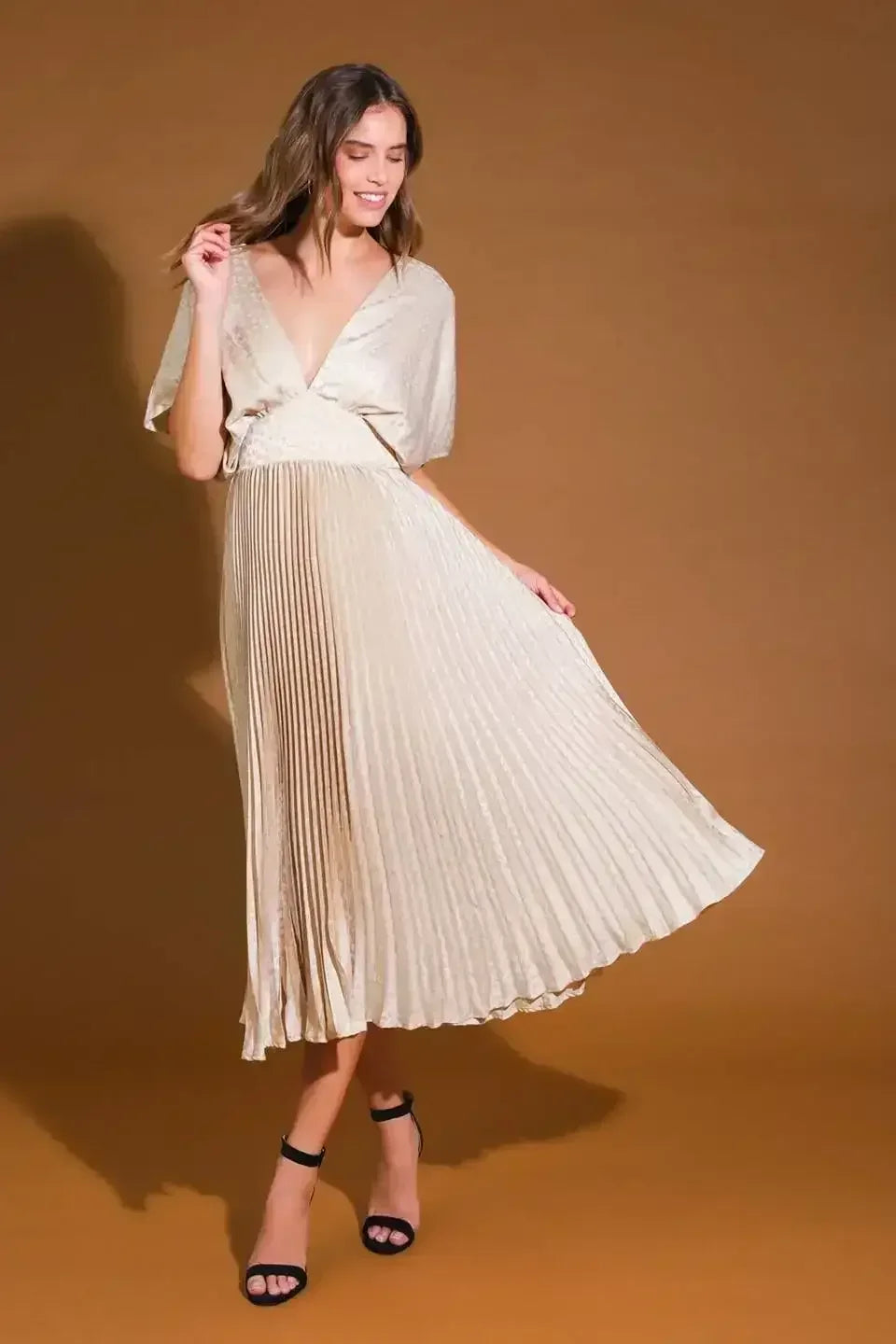 A woman in a light, pleated dress moves gracefully, smiling while holding the hem with one hand, set against a warm, brown backdrop. Black high heels complete her elegant look.