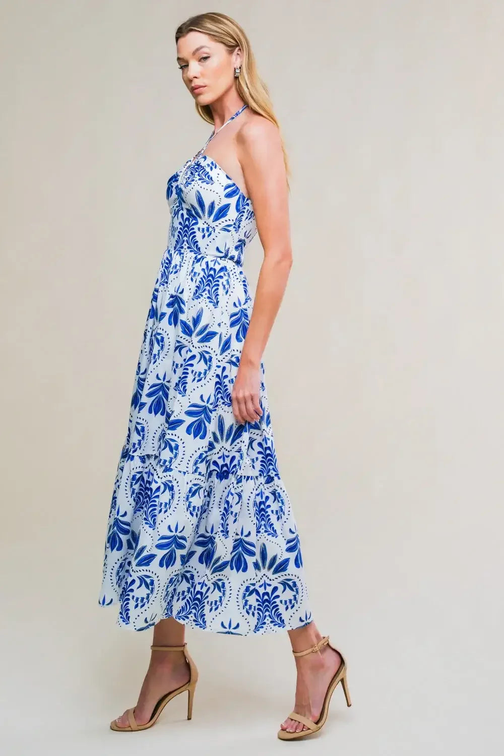 A woman stands elegantly in a long, patterned dress featuring blue floral designs against a white background. She wears nude high-heeled sandals and poses with a confident expression. The background is simple and light.