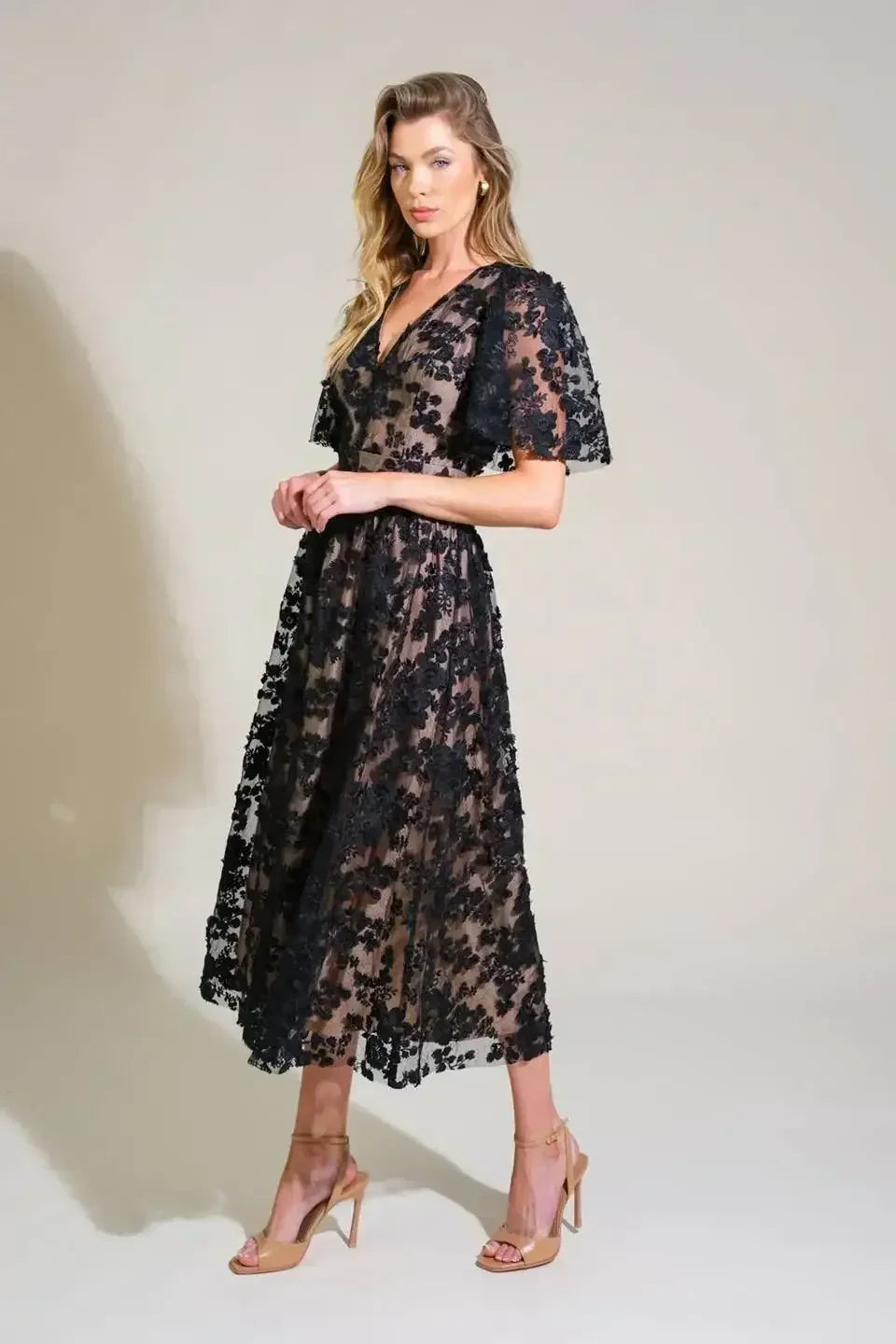A woman stands elegantly in a sheer black floral dress, posing confidently. She wears nude high-heeled shoes, with a soft, neutral backdrop enhancing the outfit's delicate, intricate details.