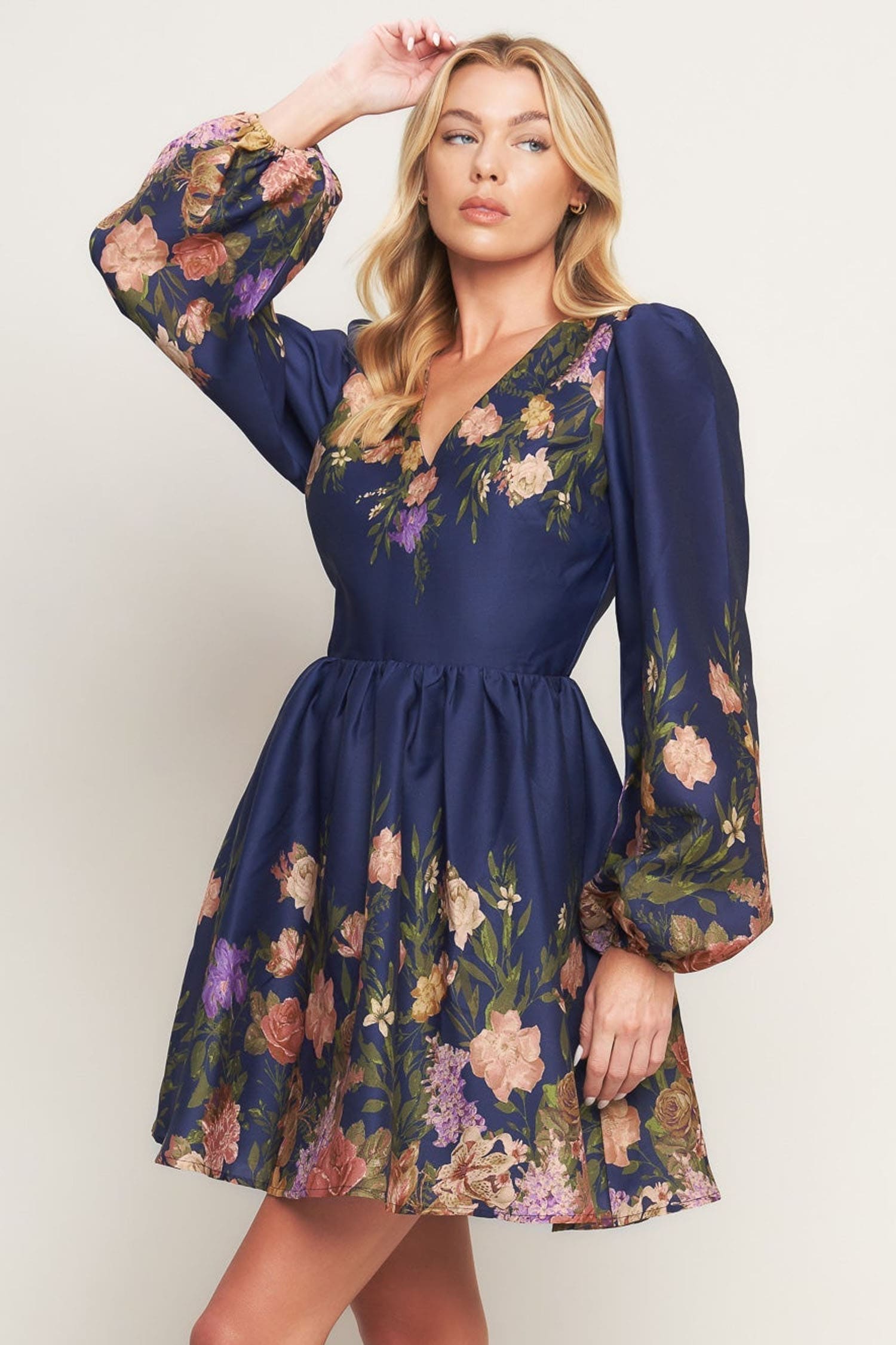 A woman modeling a navy blue mini dress with a vibrant floral print, featuring long puff sleeves and a V-neckline, set against a plain light beige background.