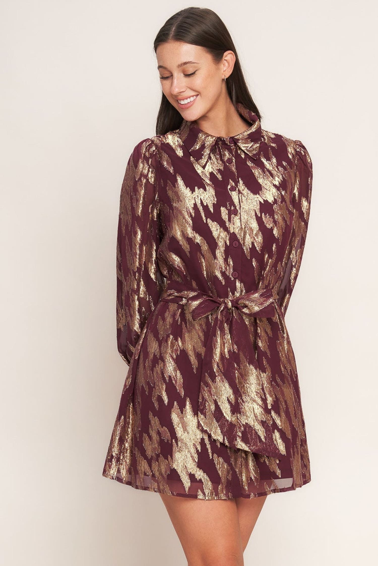 A woman modeling a long-sleeve mini dress with a metallic gold and deep burgundy abstract pattern, featuring a high neck and a self-tie waist belt, set against a plain light beige background.
