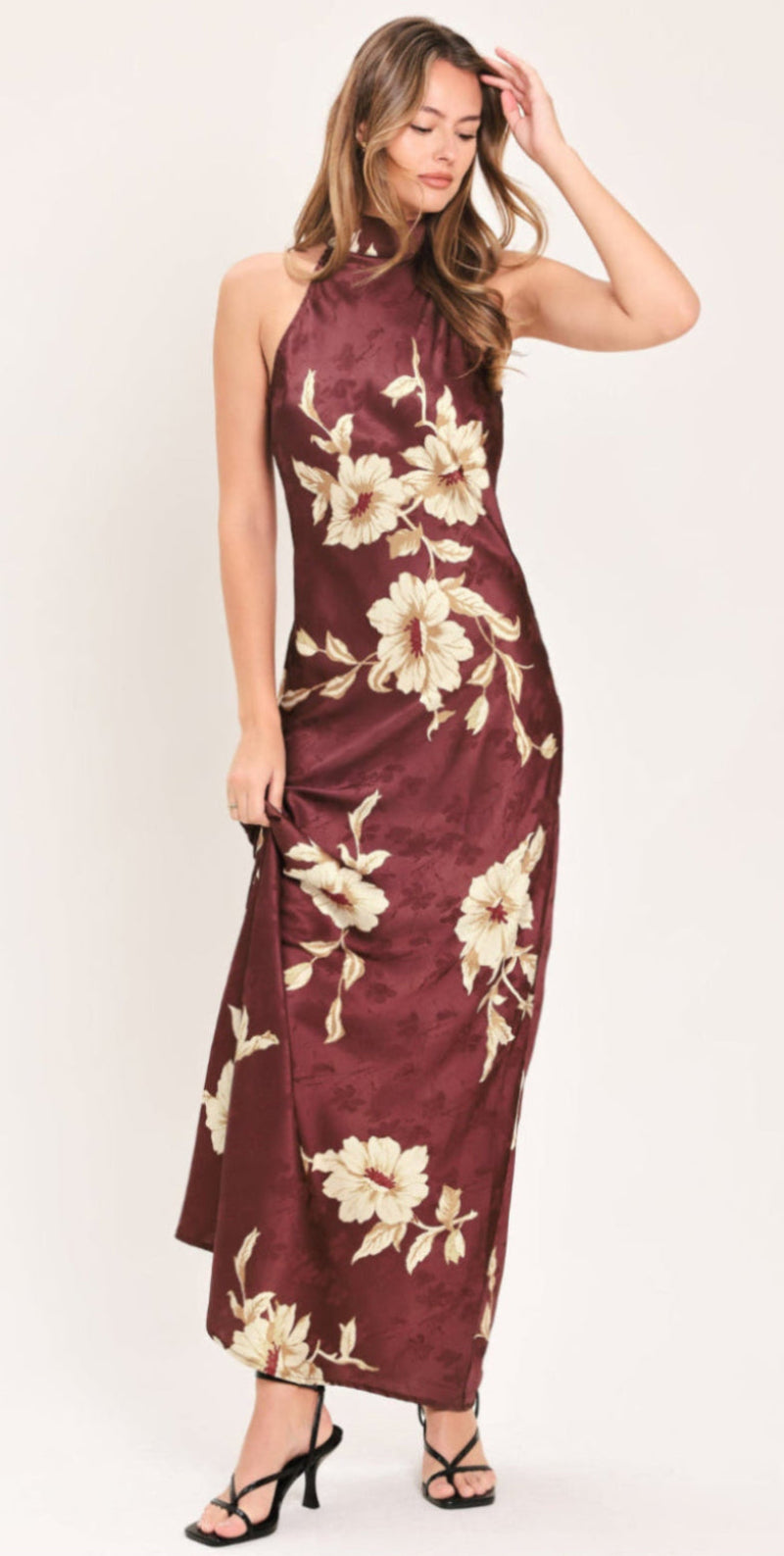 BORDEAUX IVORY BLOOM BURGUNDY MAXI DRESS | Lady Black Tie | A woman modeling a sleeveless halter-neck maxi dress in a deep burgundy satin fabric with a cream floral print, paired with black strappy high-heeled sandals against a plain off-white studio background.