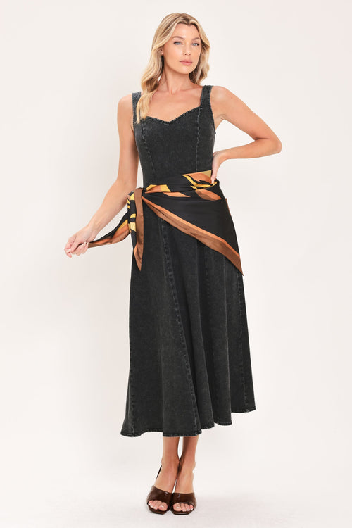 DARKWASH SCARF ELEGANCE DENIM MIDI DRESS | Lady Black Tie | A woman modeling a sleeveless black midi dress with a draped wrap-style belt featuring a brown and gold geometric pattern, set against a plain white studio background.