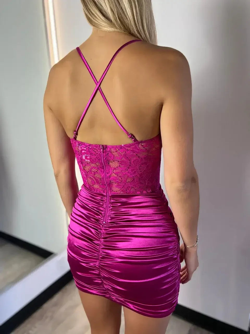 A fitted, bright pink dress with a lace top and ruched design is worn by a person seen from the back, standing against a neutral-colored wall and mirrored surface.