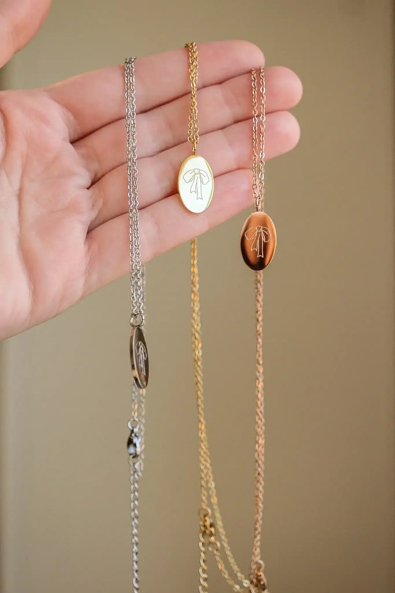 A hand holds three necklaces in silver, gold, and rose gold with oval pendants featuring a bow design, against a neutral background, showcasing the intricate details of the jewelry.