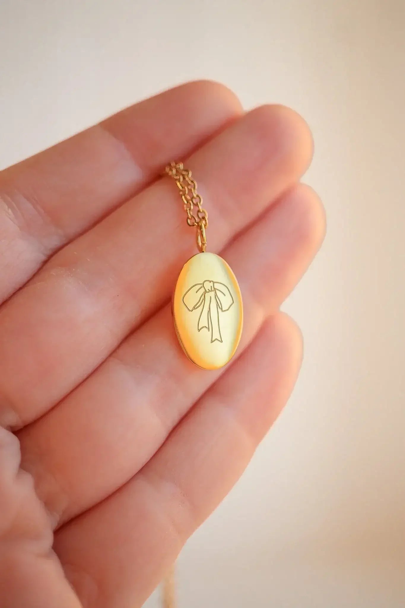 A hand holds a gold chain necklace featuring an oval pendant engraved with a ribbon design, set against a soft, neutral background that suggests indoor lighting.