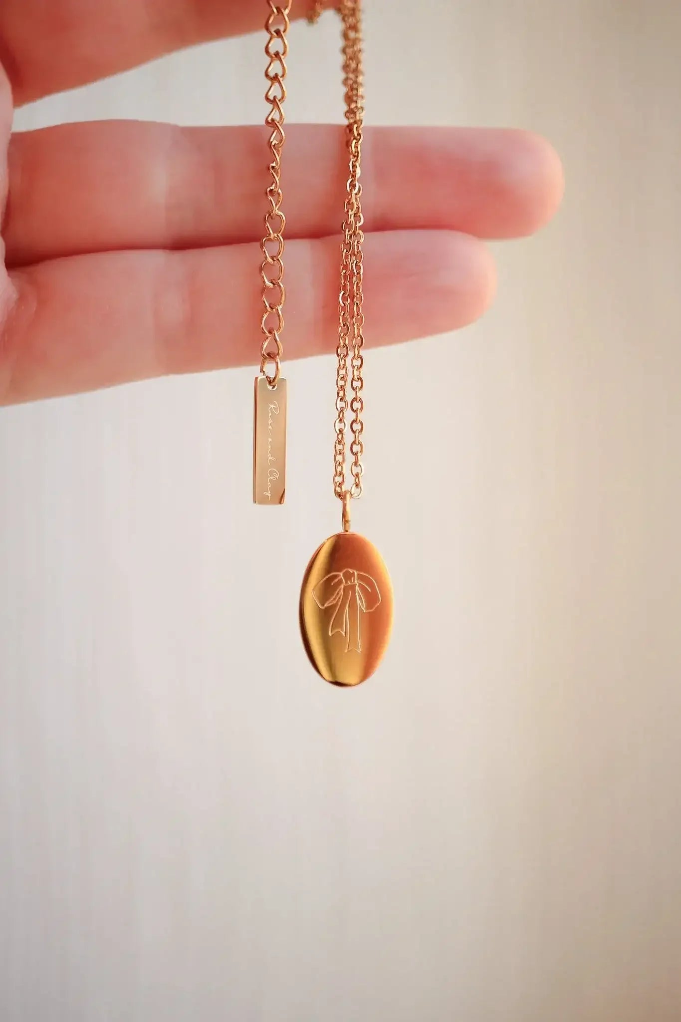 A hand holds a delicate rose gold necklace featuring an oval pendant engraved with a ribbon design and a rectangular charm inscribed with "Returned Clay," set against a light background.