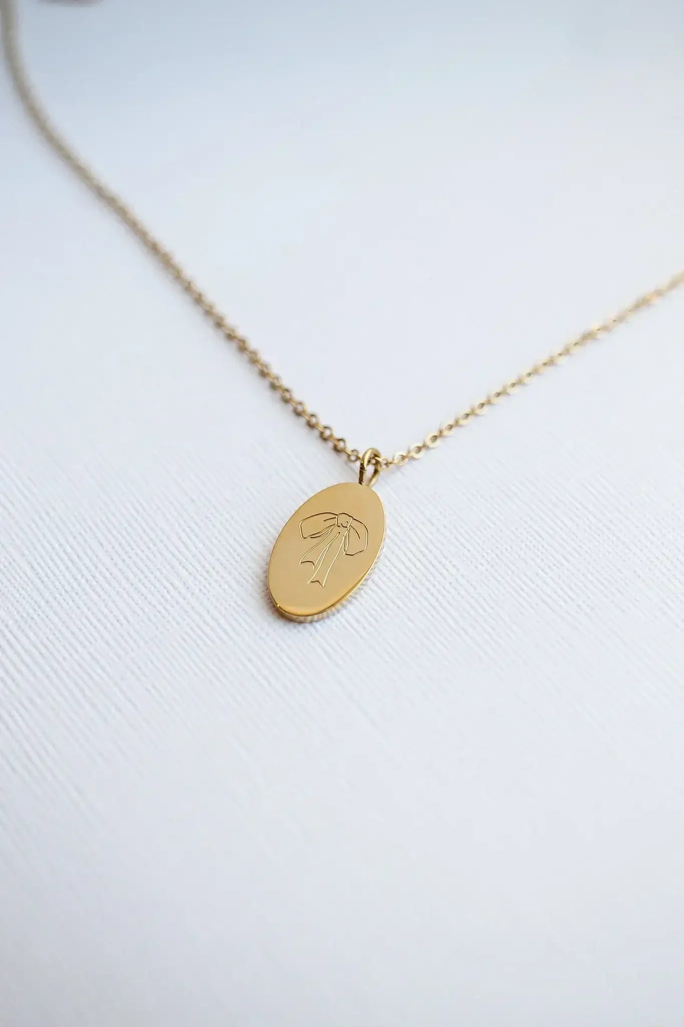 A gold necklace with an oval pendant lies on a textured white surface. The pendant features an etched design of a bow, reflecting a simple yet elegant style.