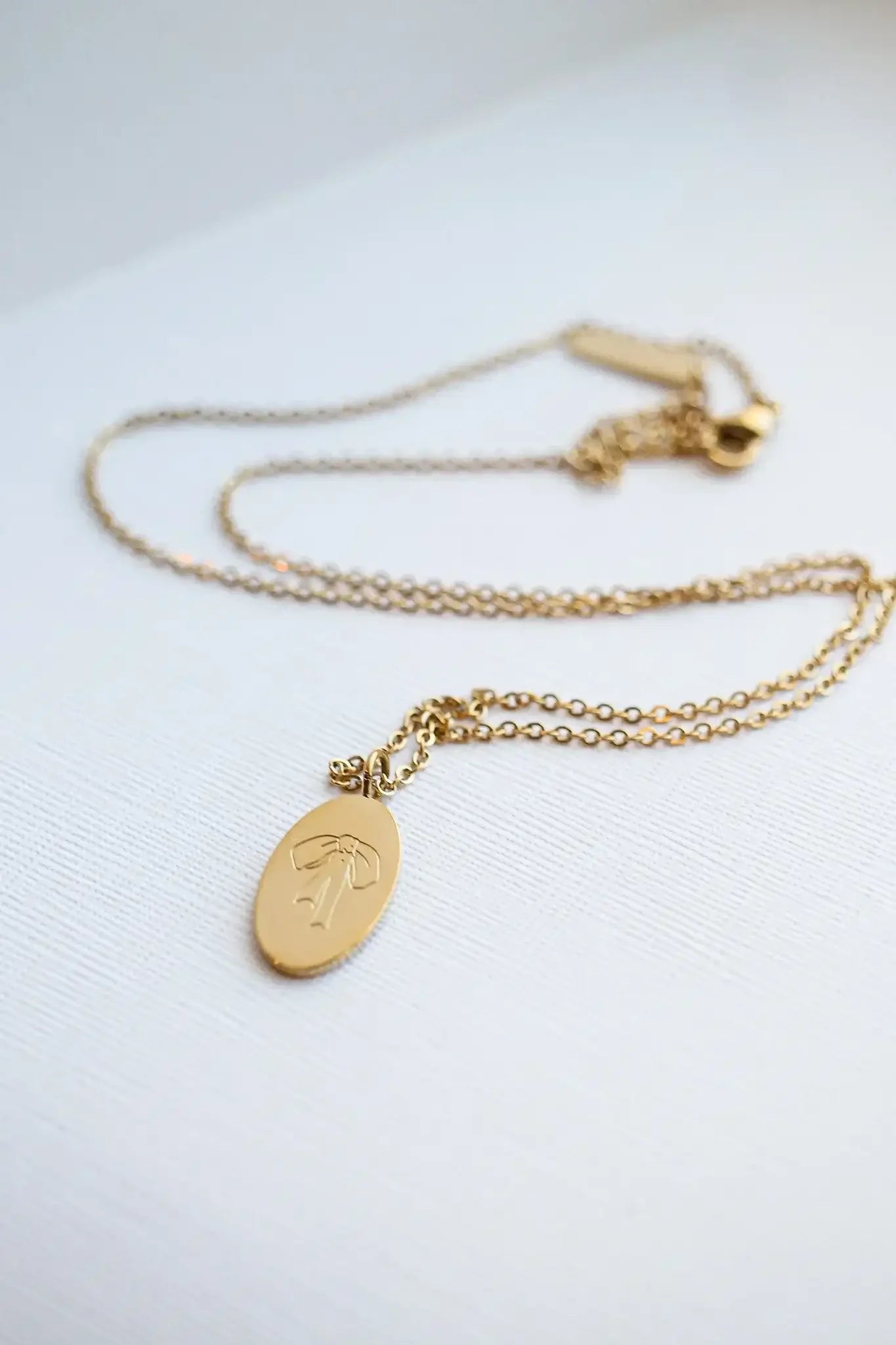 A gold necklace features an oval pendant with a bow design. The chain lies flat against a textured white background, highlighting its elegance and simplicity.