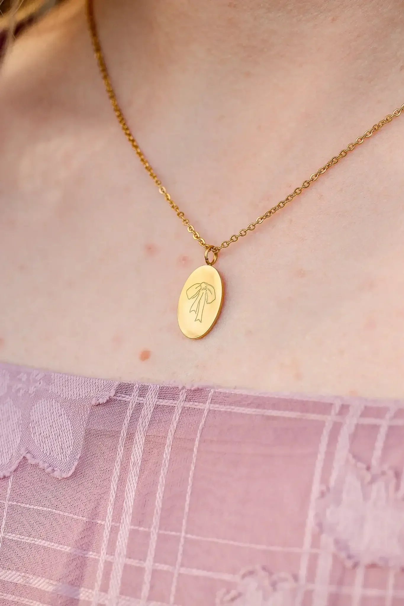A gold necklace with an oval pendant featuring a ribbon design rests on a person's neck, complementing their pink, textured clothing in a softly lit environment.