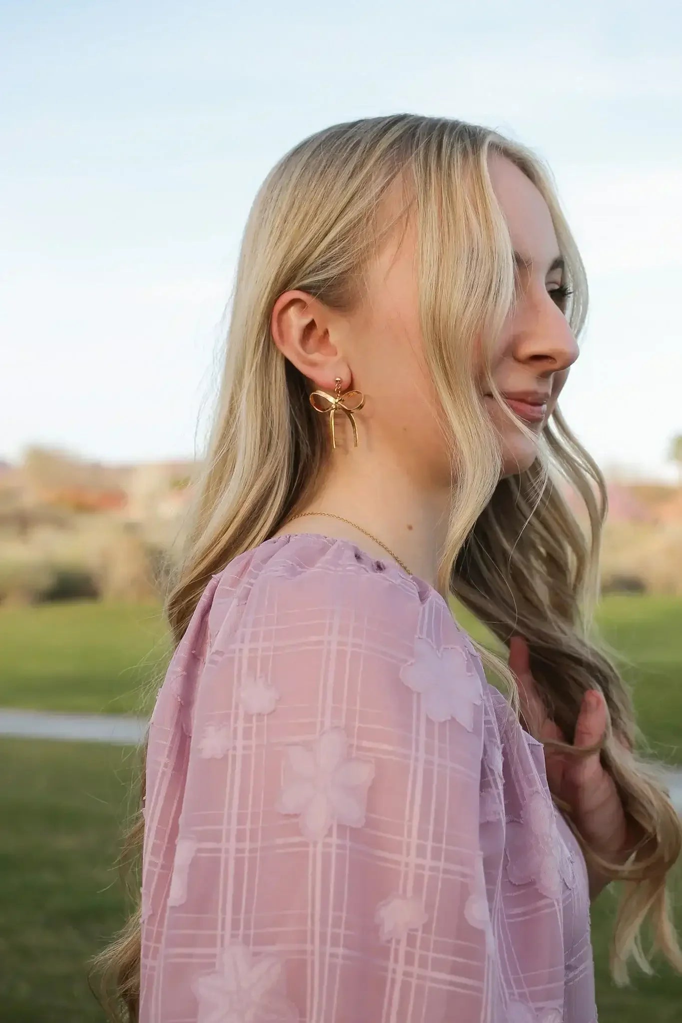 A woman with long, wavy blonde hair wears gold bow-shaped earrings, smiling softly while gently touching her hair. She stands outdoors in a green park under a clear blue sky.