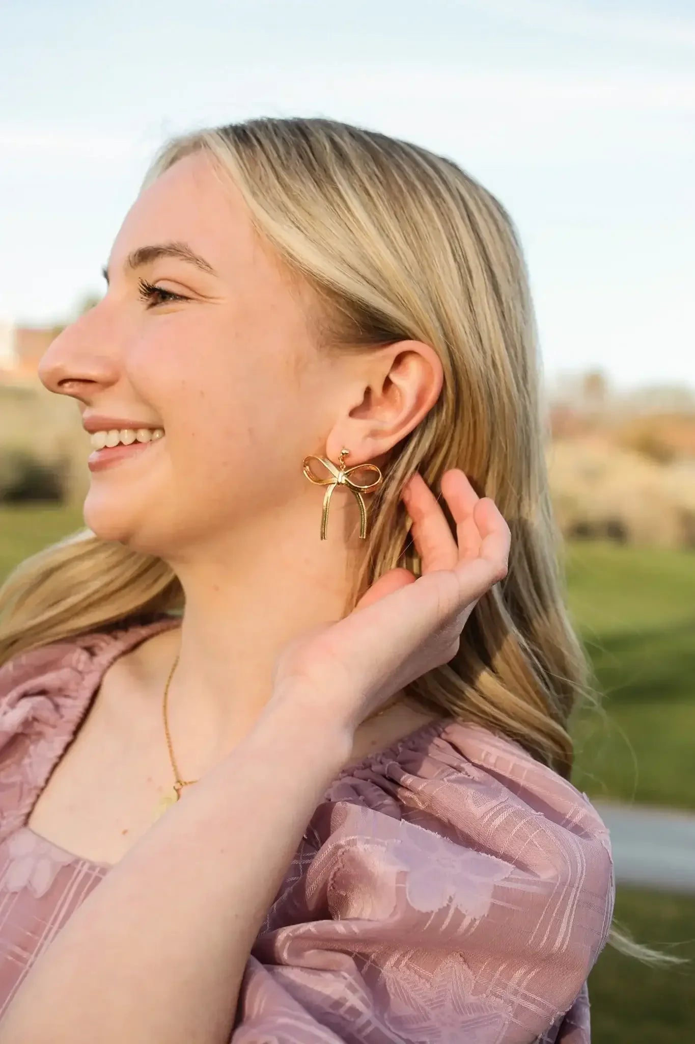 A woman with long, blonde hair smiles while gently touching her ear adorned with large, bow-shaped gold earrings. She is outdoors in a green, grassy area.