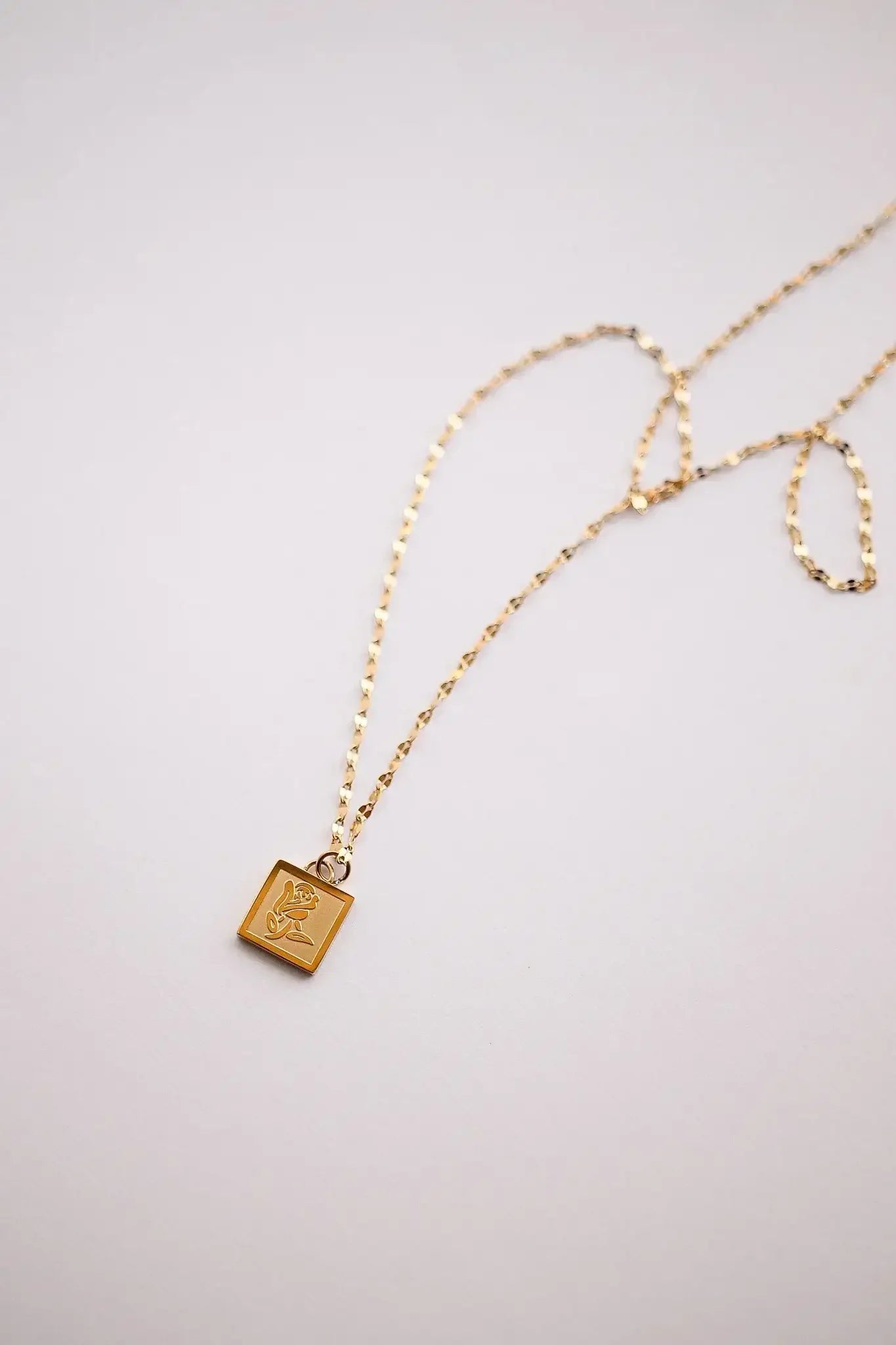 A gold necklace with a rectangular pendant showcasing an engraved design, featuring a flower and a bell. It rests against a smooth, light-colored background.