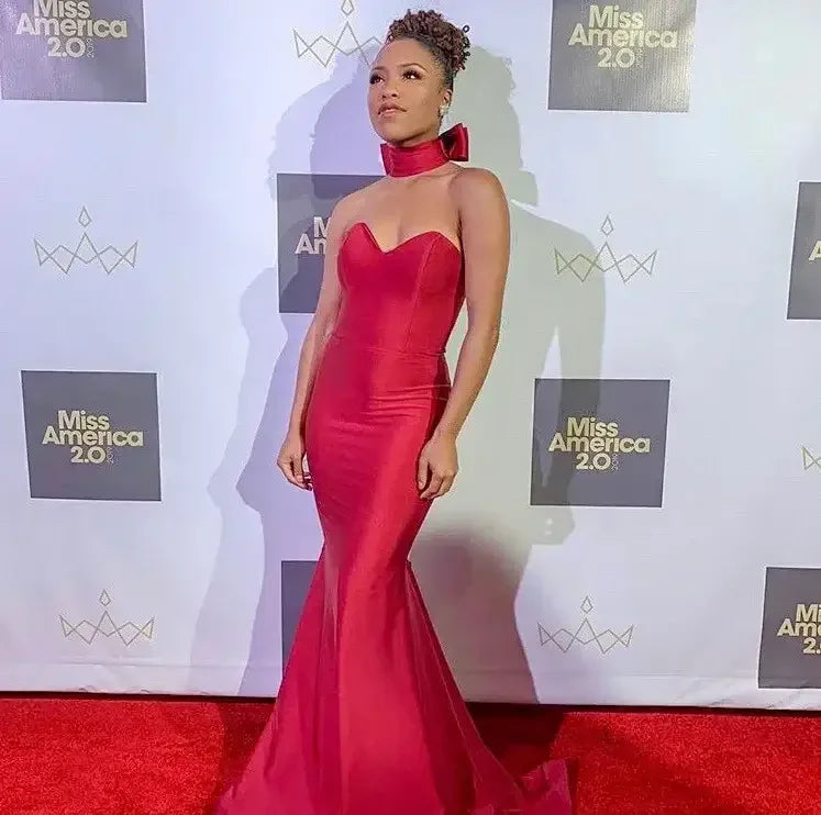 An individual stands confidently in a sleek, form-fitting red gown, accompanied by a matching choker. The backdrop features logos for "Miss America 2.0," set against a red carpet.