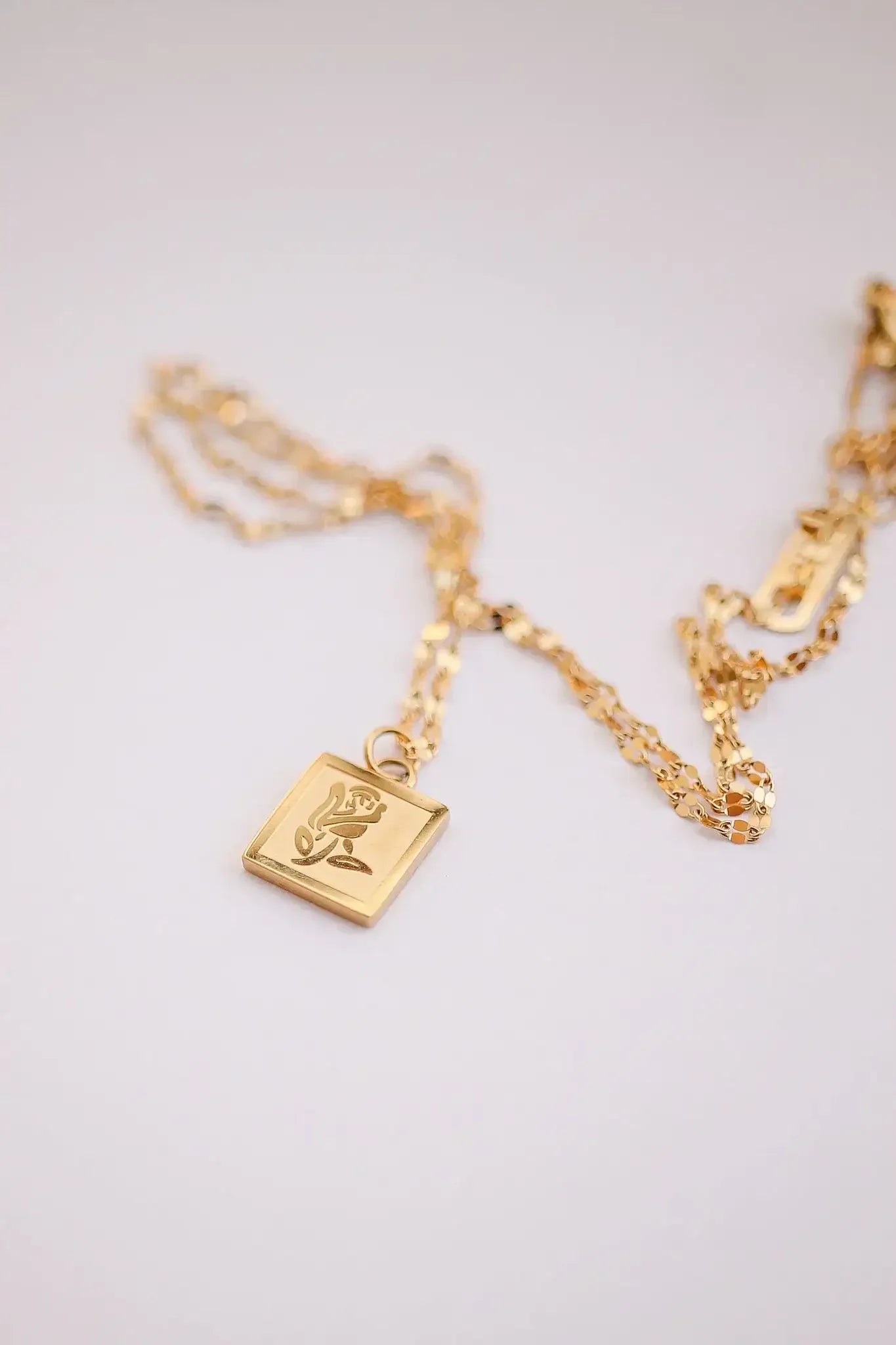 A gold necklace features a square pendant with a rose design. The chain is intricately linked, lying against a light, neutral background, highlighting its elegant appearance.