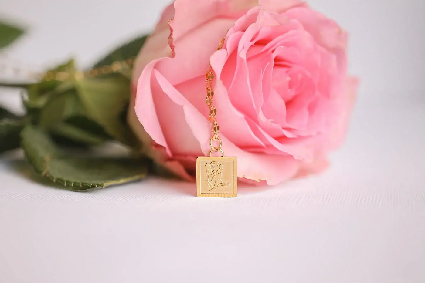 A pink rose rests on a light surface, with a delicate gold necklace featuring a square pendant adorned with an engraved rose design draped over it.