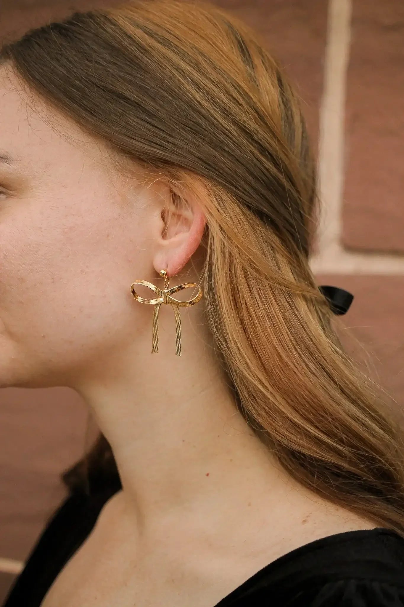 Gold earrings shaped like a bow dangle from the ear of a woman with shoulder-length hair, set against a textured, warm-colored wall, highlighting a fashion detail.