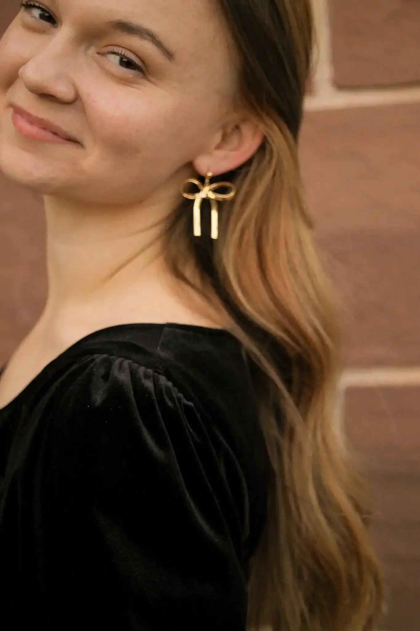 A woman with long, wavy hair is smiling slightly, wearing a black velvet shirt and oversized gold bow earrings. She is positioned against a textured brown background.