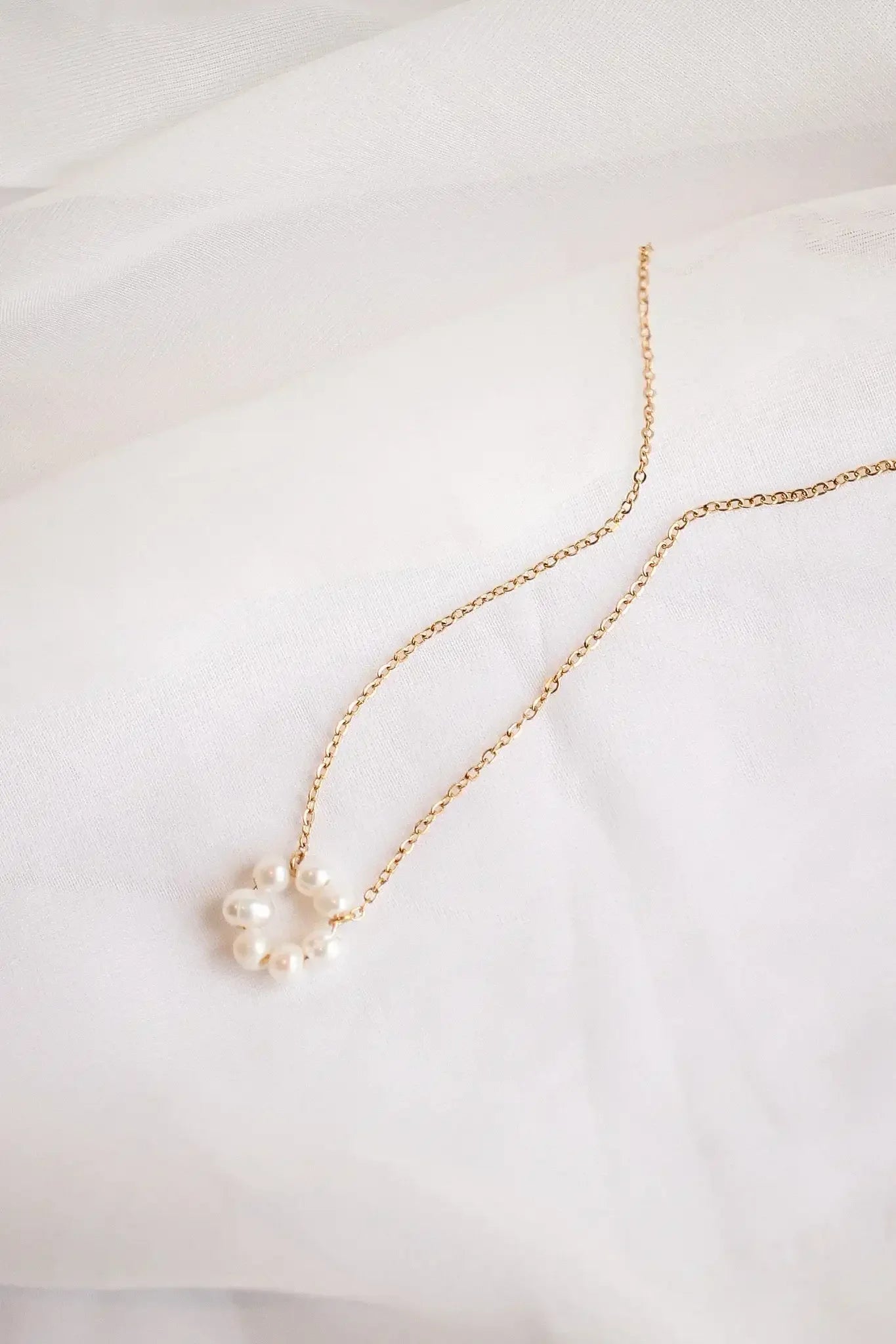 A delicate gold necklace with a cluster of small pearls rests on soft, white fabric. The pearls are arranged in a circular pattern, enhancing the necklace's elegance.