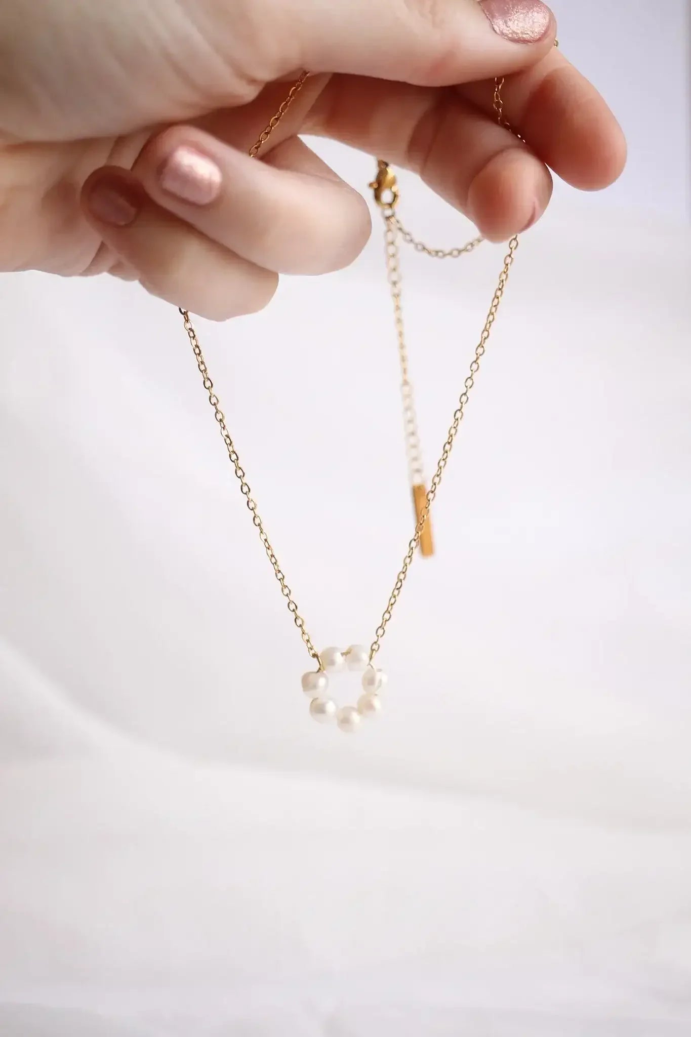 A hand holds a delicate gold chain necklace with a circular pearl pendant while set against a soft, light background. The pearls form a subtle, elegant design.