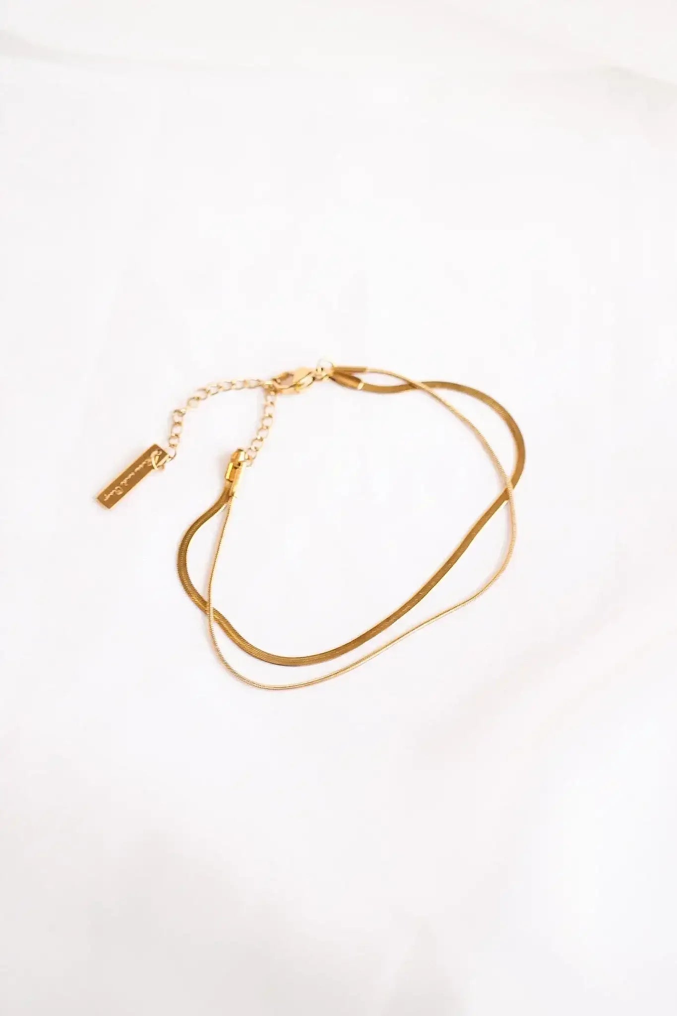 A delicate gold chain necklace rests on a soft, light-colored fabric, showcasing two slender strands and a small metal tag attached to a clasp.