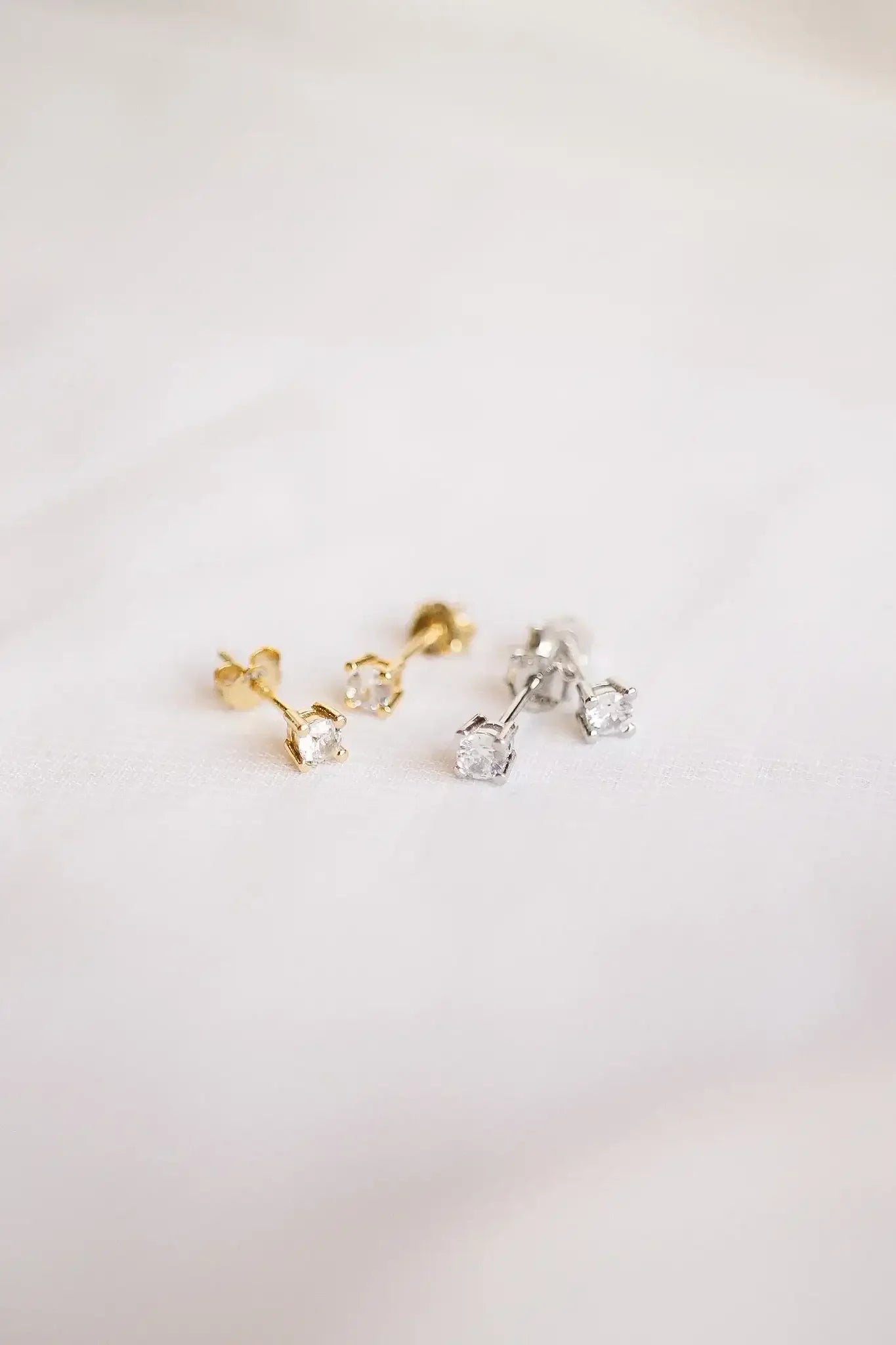 Gold and silver stud earrings, featuring small sparkling gems, rest on a light-colored textured fabric, showcasing their elegance and intricate settings in a soft, diffused light.