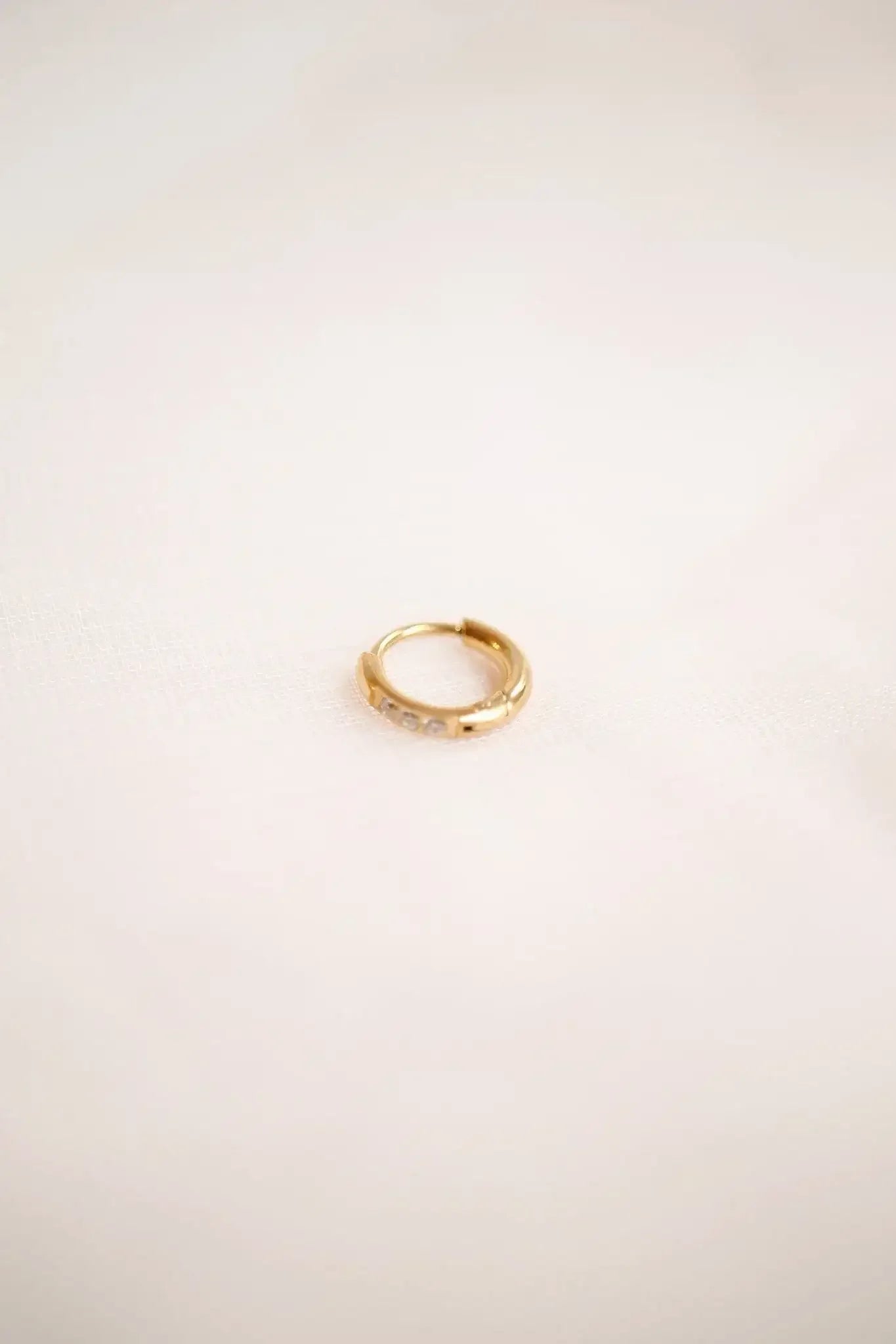 A small gold hoop earring with a smooth surface and three clear stones rests on a soft, light-colored fabric background, highlighting its elegant design and shine.