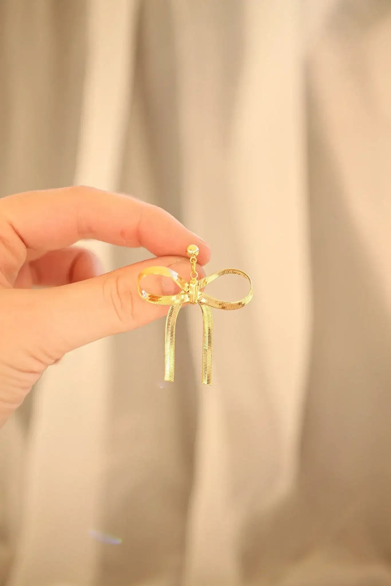 A hand holds a shiny gold bow-shaped earring, showcasing its intricate design and decorative ribbon ends against a soft, neutral background.