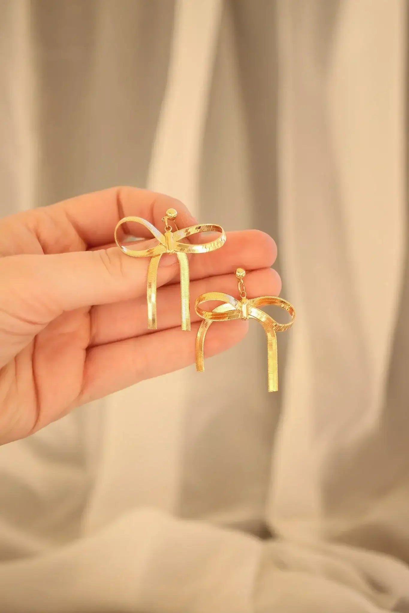 A hand holds two pairs of gold bow-shaped earrings, showcasing their decorative ribbons and clasps against a softly draped, neutral-colored background.