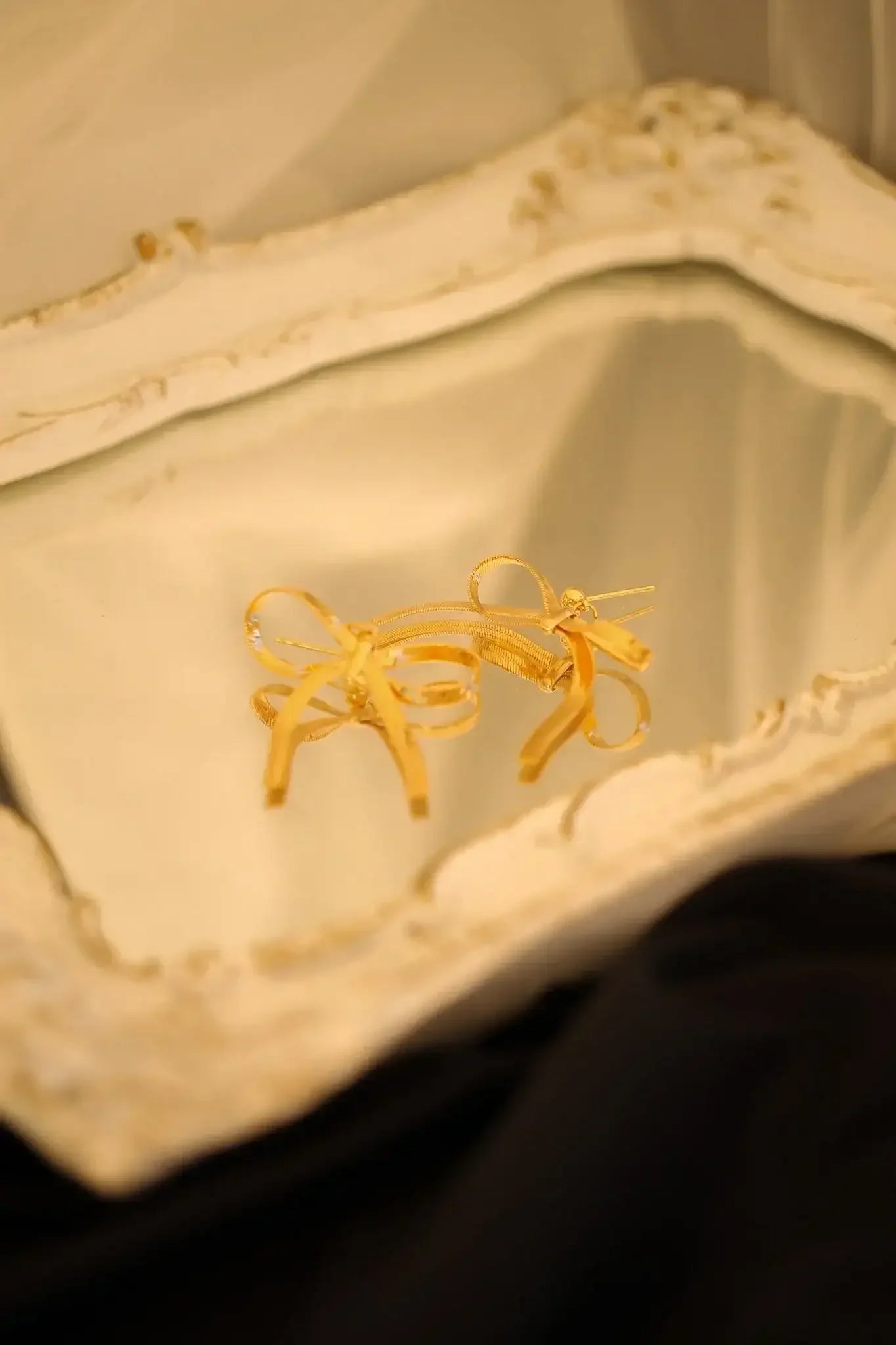 Golden earrings shaped like bows rest on an ornate, reflective surface, surrounded by soft, muted fabrics that create a delicate, elegant atmosphere. The setting enhances the jewelry's appeal.