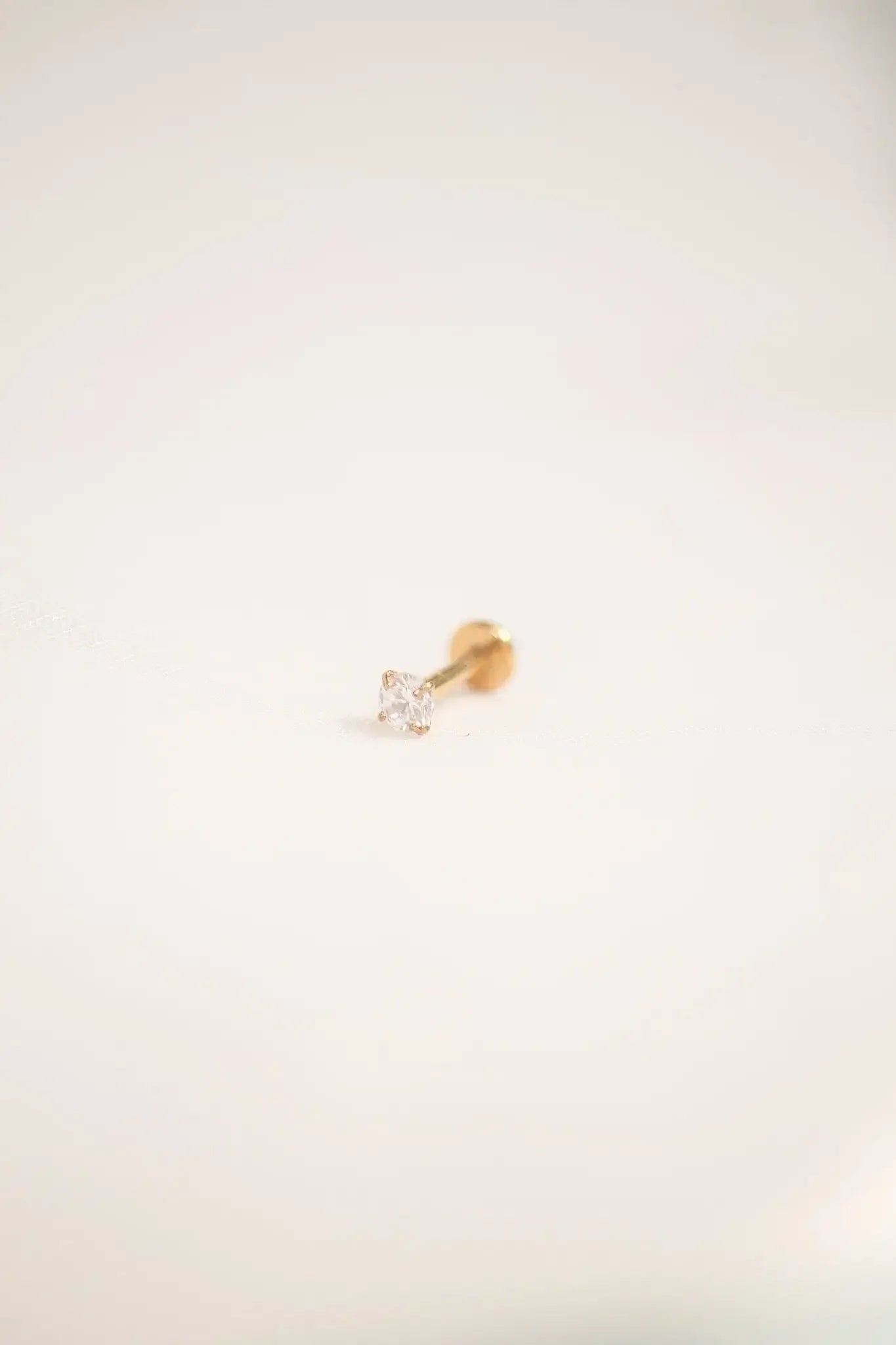 A small, golden stud earring with a sparkling clear gemstone sits flat on a soft, off-white surface, highlighting its delicate design against the neutral backdrop.