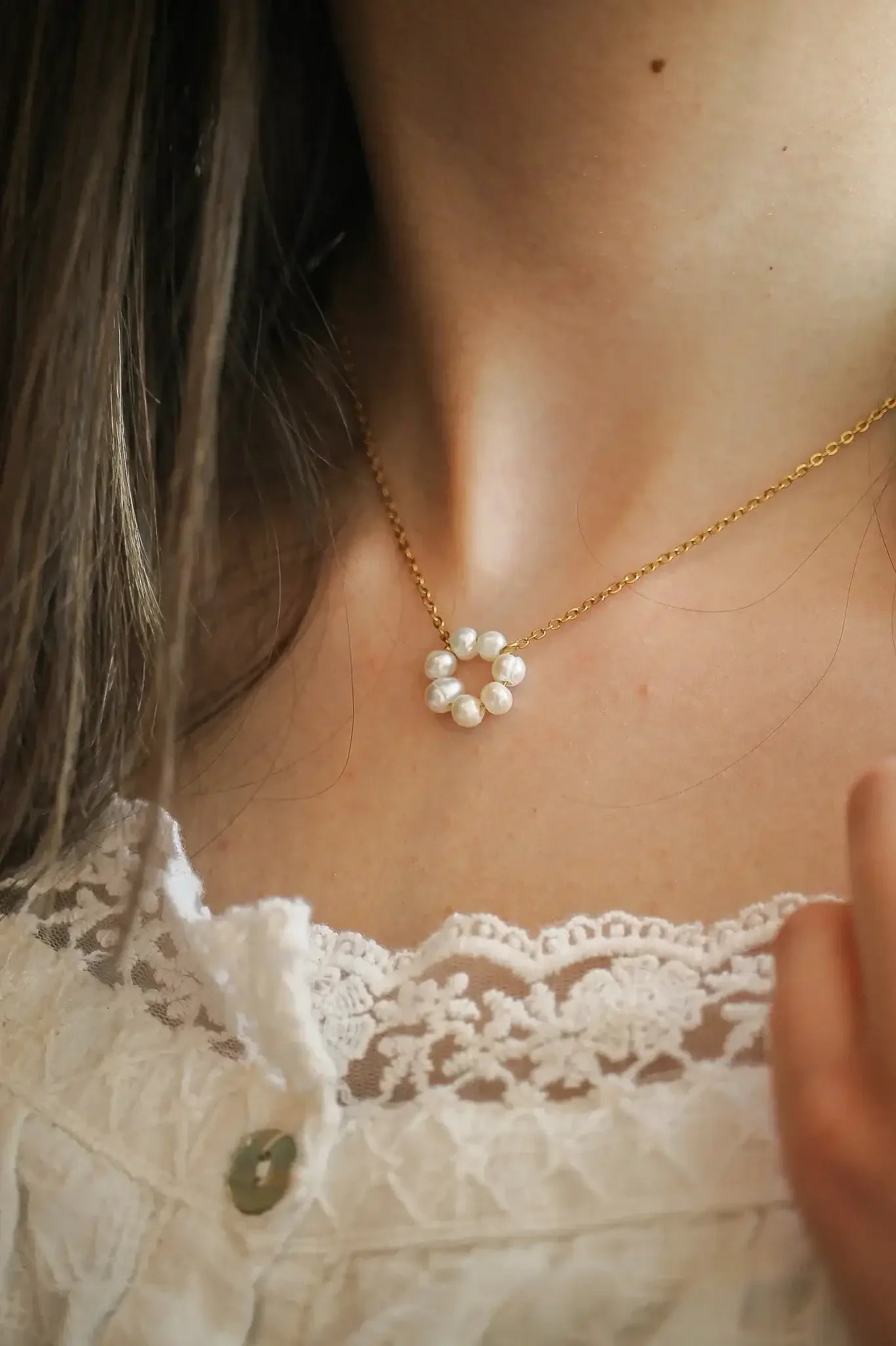 A delicate gold necklace with pearl accents sits around a neck, complementing a white lace top. The wearer gently touches the necklace, highlighting its elegant design.