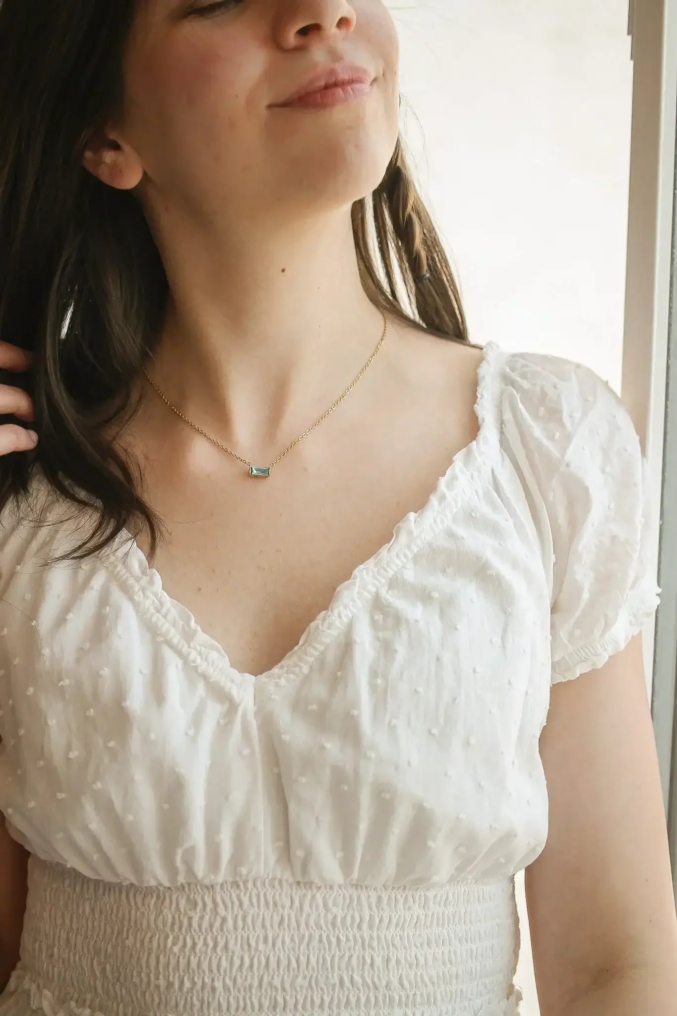 A young woman in a white, textured blouse smiles softly while playing with her hair. She wears a delicate gold necklace with a rectangular, shimmering pendant, set against a light background.