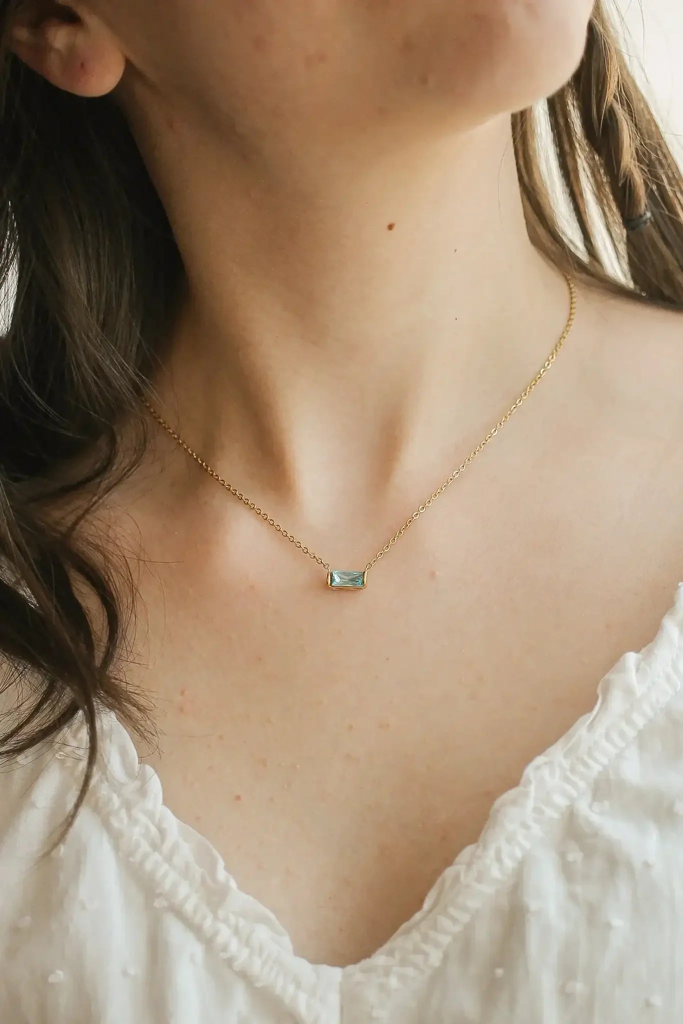A delicate gold necklace with a rectangular gemstone pendant rests on the smooth skin of a person's neck, complementing their simple white blouse. The background is softly blurred.