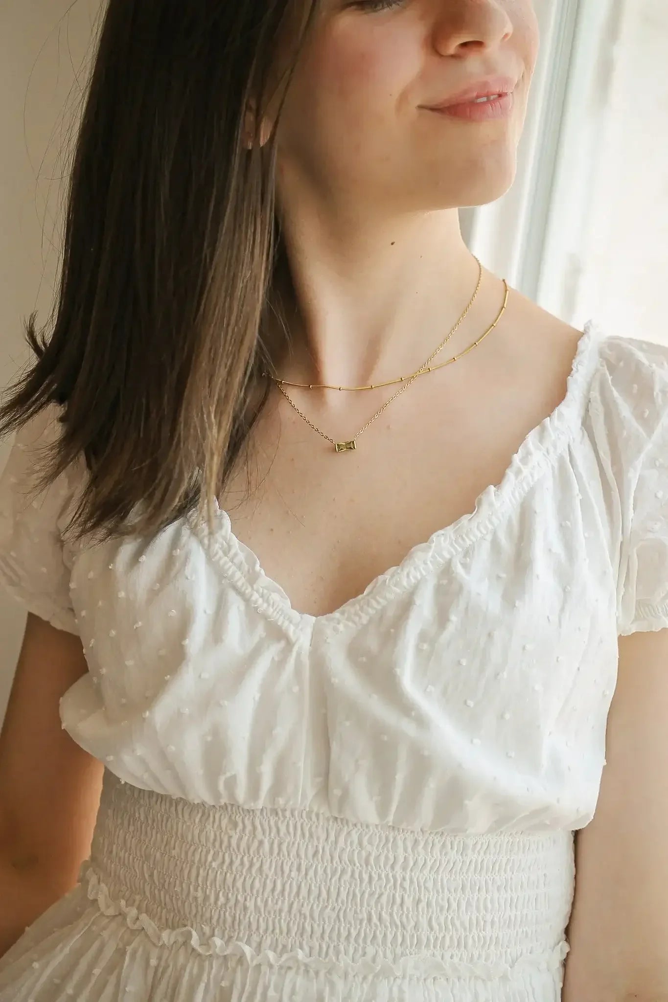 A young woman with shoulder-length hair smiles, wearing a white, textured dress. She showcases layered gold necklaces, standing near a window that lets in soft natural light.
