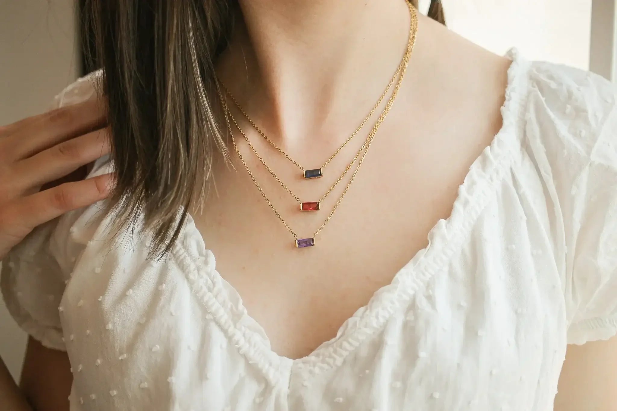 A person wears three layered gold necklaces with colorful rectangular pendants. One hand touches their neck. The outfit features a white, textured blouse and the background appears softly blurred.
