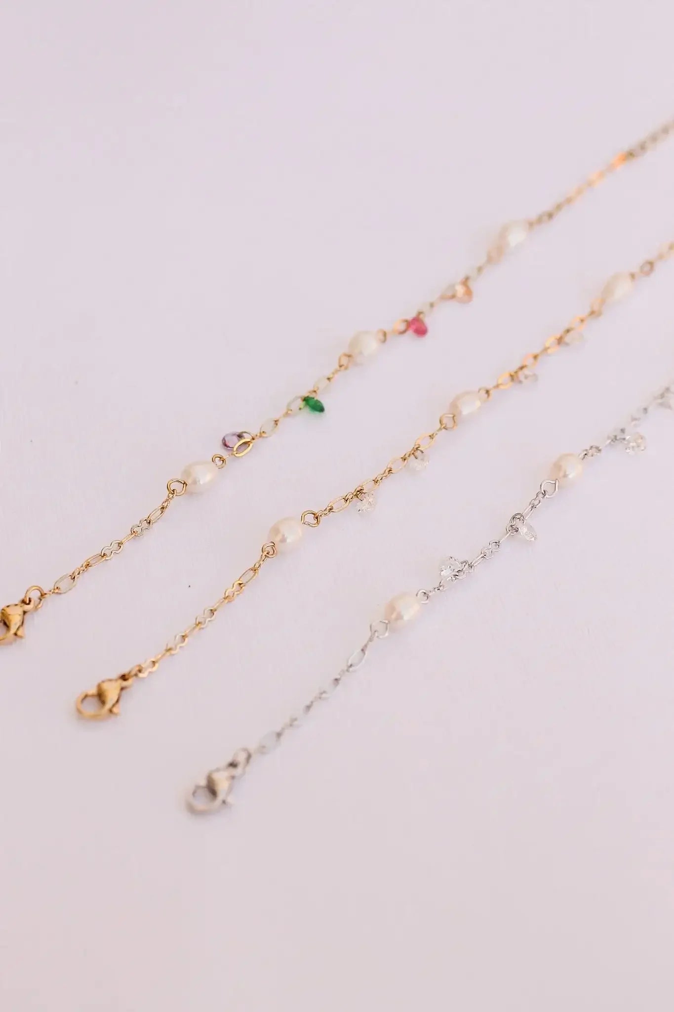 Three delicate chains composed of gold and silver are adorned with pearls and colorful gemstones. They lie flat on a light-colored surface, showcasing their elegant designs and clasps.