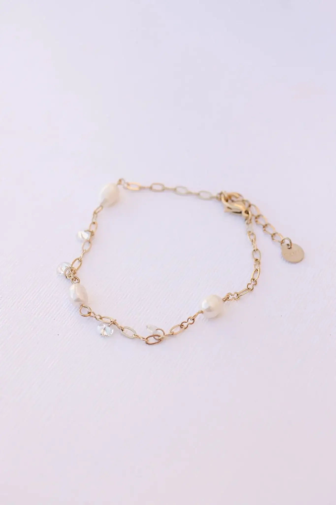 A delicate gold bracelet features lustrous pearls and small crystals linked together. It rests flat on a textured white surface, showcasing its elegant design and materials.