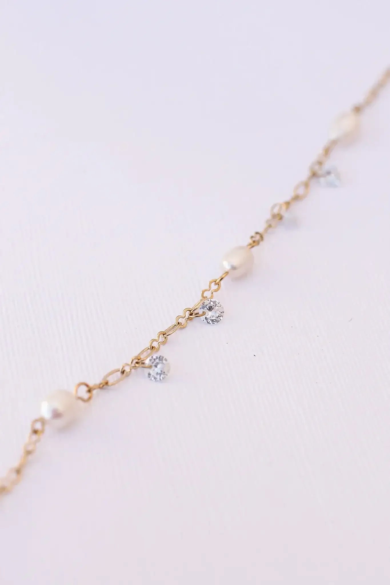 A delicate gold chain features alternating pearls and sparkling rhinestones, arranged symmetrically. The chain rests gently against a soft, textured white background, highlighting its elegance.