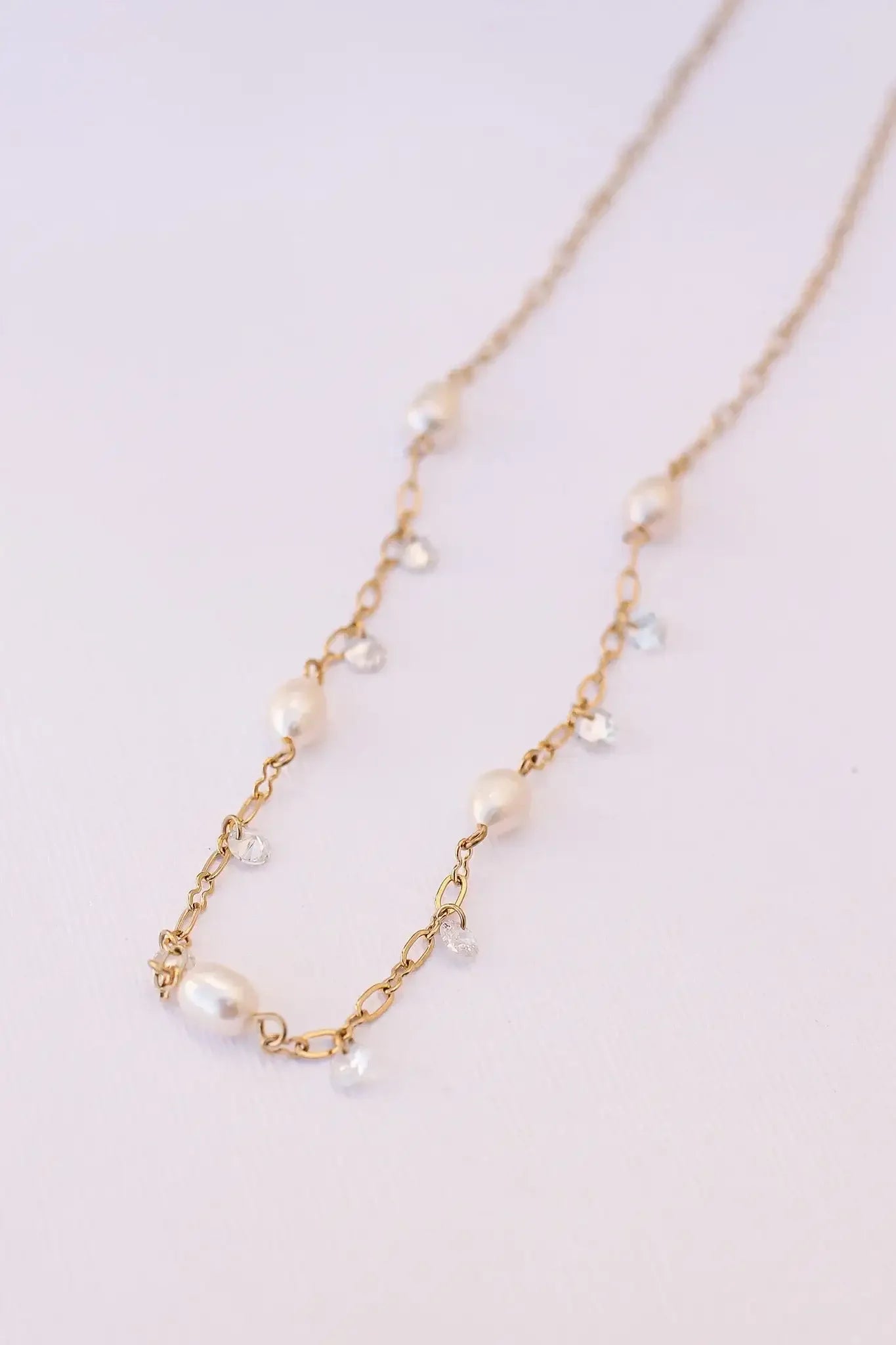 A gold chain necklace featuring alternating white pearls and sparkling crystals is elegantly laid out against a soft, textured white background. The design is delicate and sophisticated.