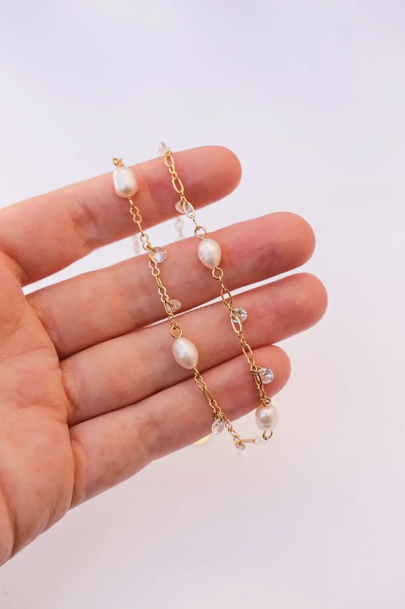 A hand holds a delicate gold chain adorned with irregularly shaped pearls and small crystals, elegantly displaying the jewelry against a soft, light-colored background.
