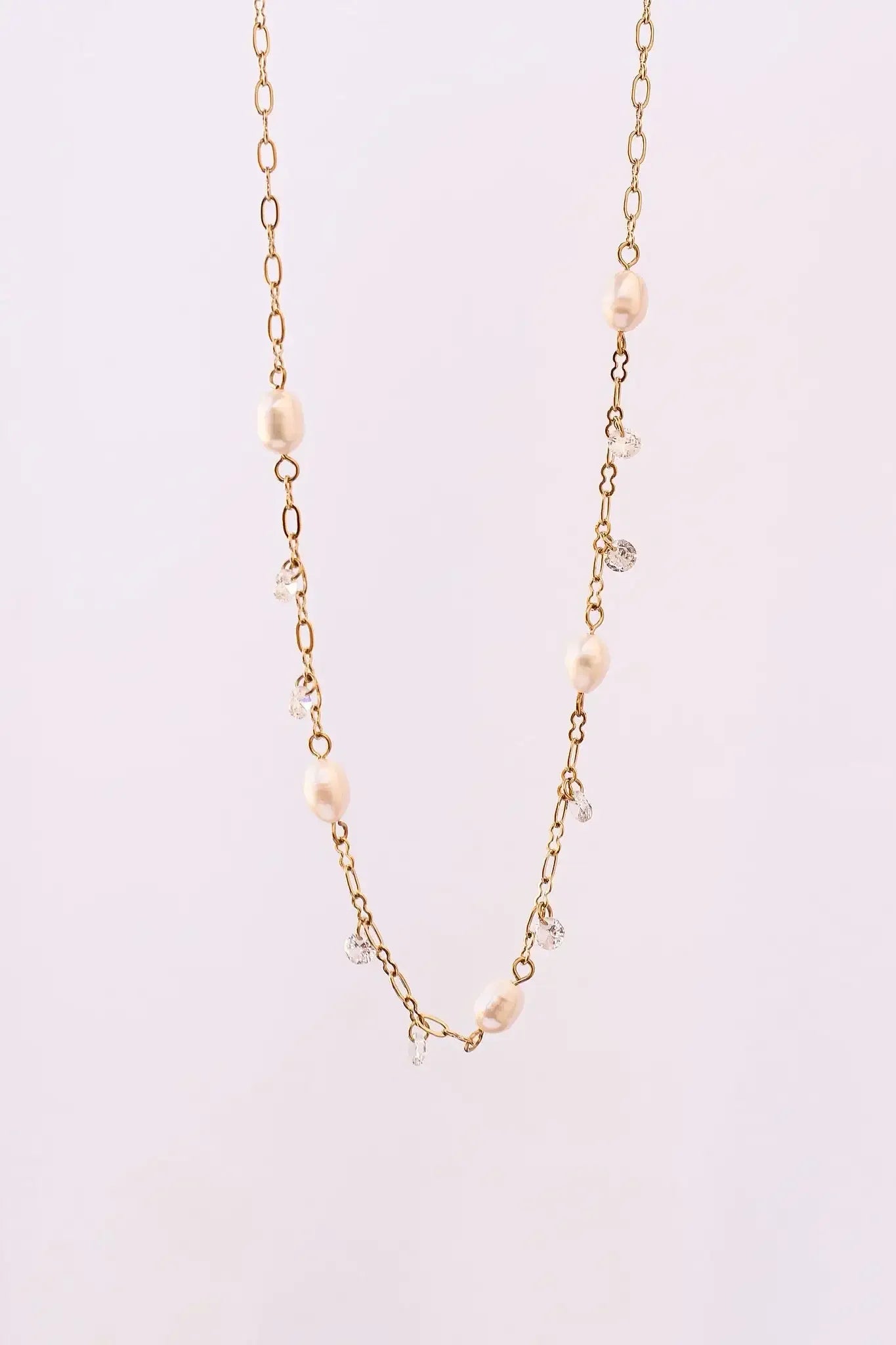 A delicate gold chain necklace adorned with alternating freshwater pearls and clear crystal beads hangs against a soft white background, showcasing its elegant and refined design.